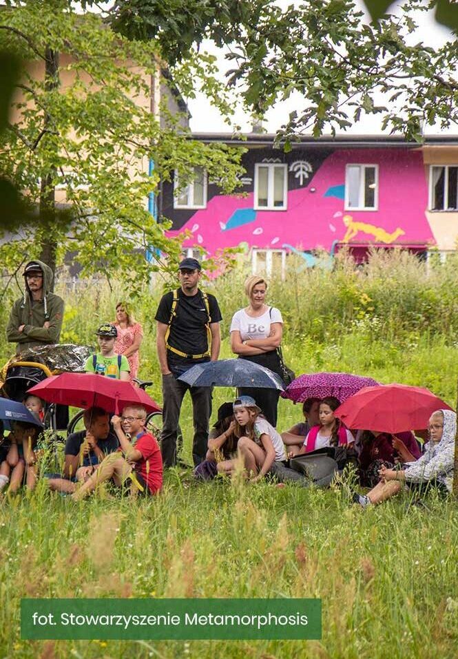 Dzieci siedzące na zielonej trawie pod parasolami. W tle budynek Hajnówka Centralna-Stacja Kultury.