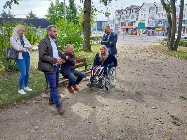 Grupa osób rozmawiająca w parku. Dwóch mężczyzn i kobieta stoją, uśmiechają się. Mężczyzna siedzi na ławce i rozmawia z kobietą siedzącą na wózku inwalidzkim.