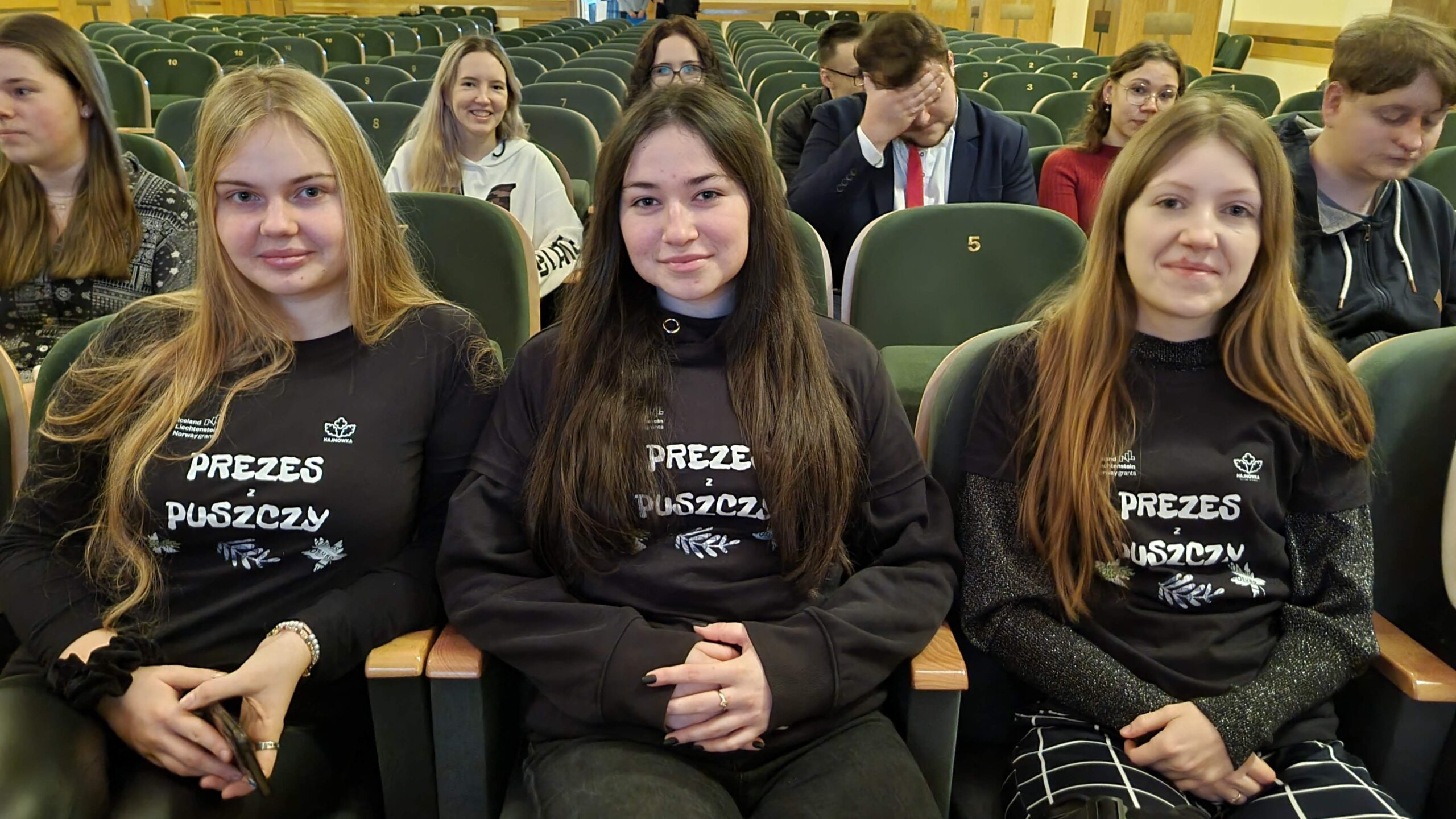 Trzy młode dziewczyny siedzą uśmiechnięte w pierwszym rzędzie sali widowiskowej.